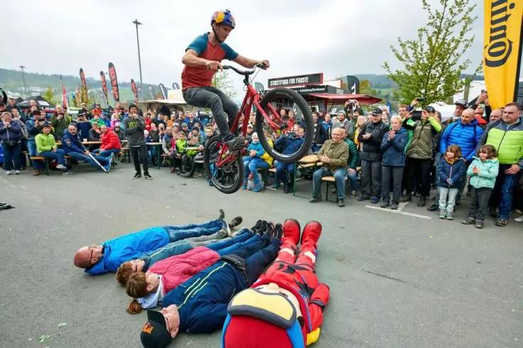 Person auf Fahrrad in der Luft &uuml;ber vier am Boden liegenden Personen, Zuschauer am Stra&szlig;enrand, Fahnen und B&auml;ume im Hintergrund