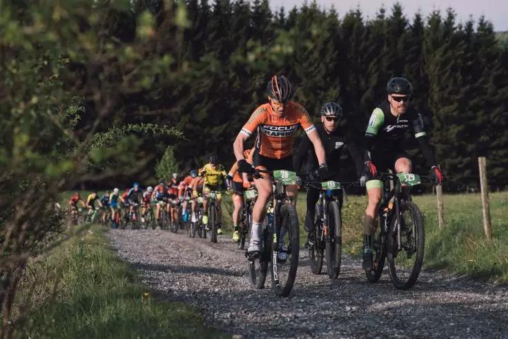 Mehrere Radfahrer mit Helmen und Startnummern fahren auf einem Schotterweg, Gras an den Seiten, B&auml;ume im Hintergrund