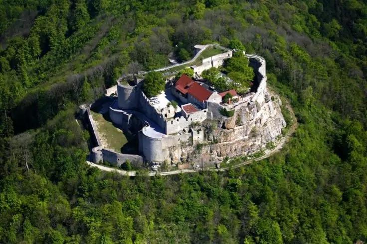 Luftaufnahme eines steinernen Geb&auml;udekomplexes mit umlaufenden Mauern und roten D&auml;chern auf einem Felsplateau, umgeben von dichtem gr&uuml;nem Wald