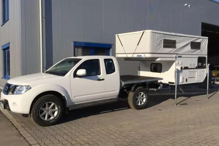 Wei&szlig;er Pickup mit Doppelkabine und aufgesetzter aufgeklappter Wohnkabine auf offenem Pritschenwagen, St&uuml;tzbeine ausgefahren, geparkt auf gepflastertem Hof neben einer grauen Industriehalle mit blauen Fensterrahmen