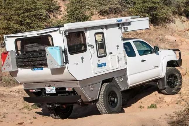 Wei&szlig;er Pickup mit aufgesetztem Camperaufbau auf sandigem Boden, trockene B&uuml;sche und felsige H&auml;nge im Hintergrund