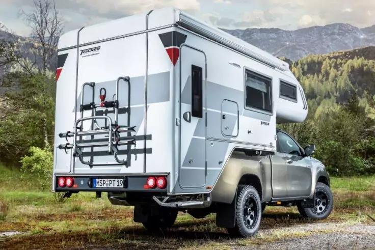 R&uuml;ckansicht eines grauen Pickups mit wei&szlig;er Wohnkabine auf der Ladefl&auml;che, montierter Fahrradtr&auml;ger am Heck, B&auml;ume und Berge im Hintergrund