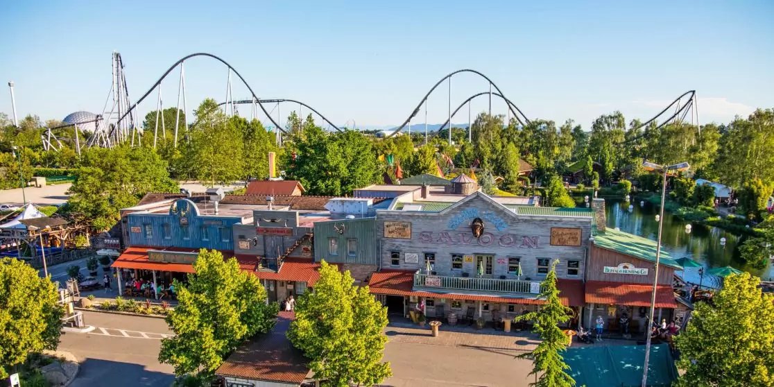 Reihe von Gesch&auml;ftsgeb&auml;uden mit orangefarbenen Markisen, B&auml;umen und parkenden Autos im Vordergrund; im Hintergrund gro&szlig;e Stahl-Achterbahn und blauer Himmel