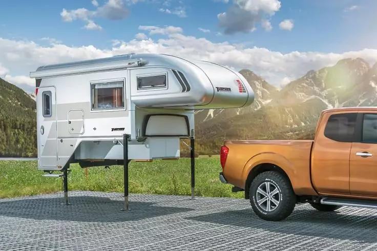 Wei&szlig;e Wohnkabine auf St&uuml;tzen neben einem orangefarbenen Pickup auf gepflastertem Platz, Berge und blauer Himmel im Hintergrund