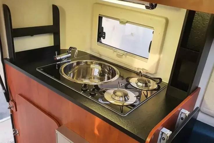 K&uuml;chenzeile mit rundem Edelstahlsp&uuml;lbecken, Zweiflamm-Gaskocher, schwarzer Arbeitsplatte, Armatur, kleinem rechteckigem Fenster und Holzschrank