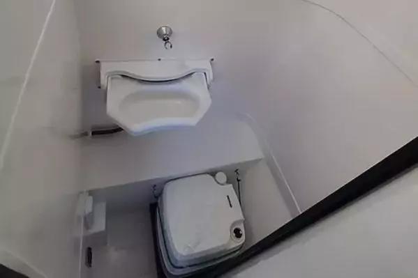 Wei&szlig;es wandmontiertes Waschbecken mit chromener Armatur oben; darunter kompakte wei&szlig;e Toilette mit geschlossenem Deckel; wei&szlig;e Innenw&auml;nde; schwarzer T&uuml;rrahmen rechts