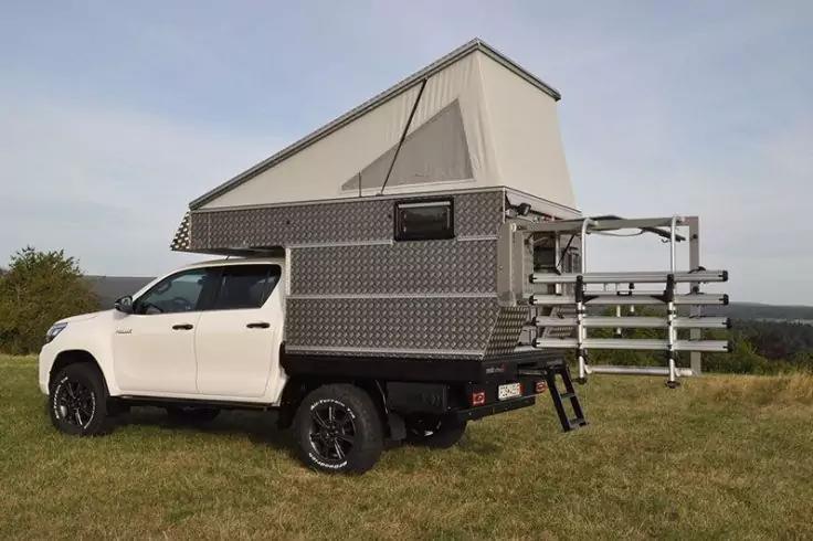Wei&szlig;er Pickup mit grauer Aufsatzkabine und aufgeklapptem Aufstelldach, offene Heckplattform mit Leiter, auf einer grasigen Fl&auml;che vor B&auml;umen und dem Horizont