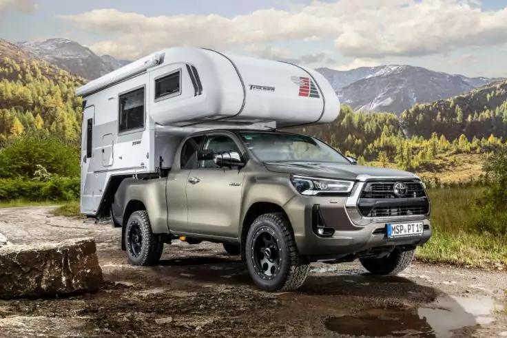 Pick-up mit aufgesetztem Alkoven-Wohnaufbau auf felsigem Untergrund; Nadelwald und Berge im Hintergrund, bew&ouml;lkter Himmel