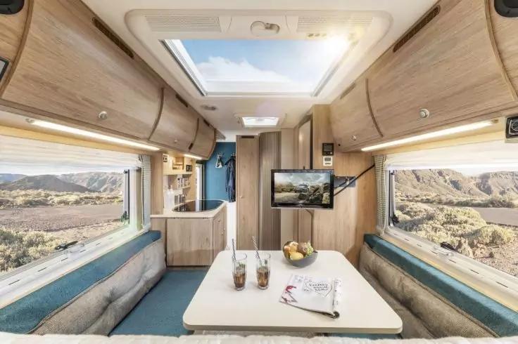 Innenraum eines Wohnmobils mit Tisch, zwei Sitzb&auml;nken, Obstschale, zwei Getr&auml;nken, offenem Dachfenster und Fenstern mit Landschaftsblick, K&uuml;chenzeile und kleinem Fernseher