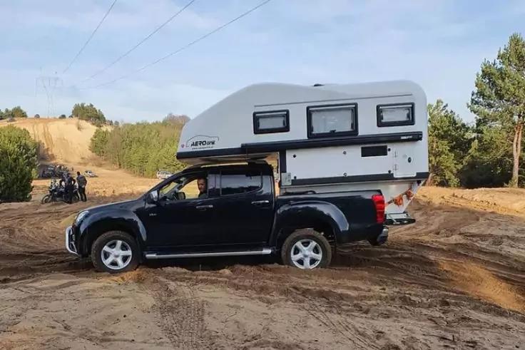 Schwarzer Pickup mit wei&szlig;em Aufbau auf der Ladefl&auml;che, drei kleine Fenster im Aufbau, sandiger Boden, Nadelb&auml;ume im Hintergrund, Stromleitungen am Himmel