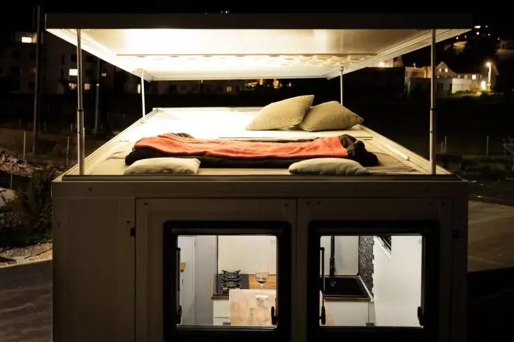 Container&auml;hnliches Geb&auml;ude bei Nacht, Dachbett mit Matratze, zwei Kissen und orangefarbener Decke unter einem Vordach; darunter zwei beleuchtete Fenster mit sichtbarer Innentreppe und M&ouml;beln