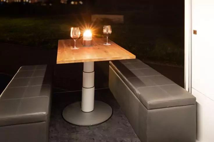 Quadratischer Holztisch mit mittigem S&auml;ulenfu&szlig;, zwei graue Polsterb&auml;nke, zwei Weingl&auml;ser und eine brennende Kerze im Glas, dunkler Hintergrund