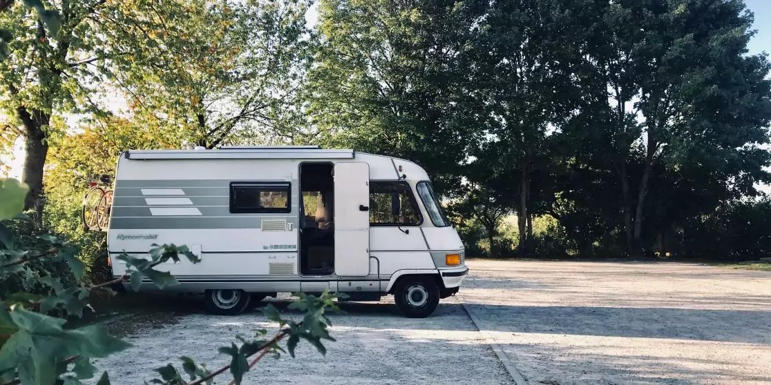 Wei&szlig;es Wohnmobil auf einem sonnigen Parkplatz neben B&auml;umen, Seitenansicht mit ge&ouml;ffneter Seitent&uuml;r