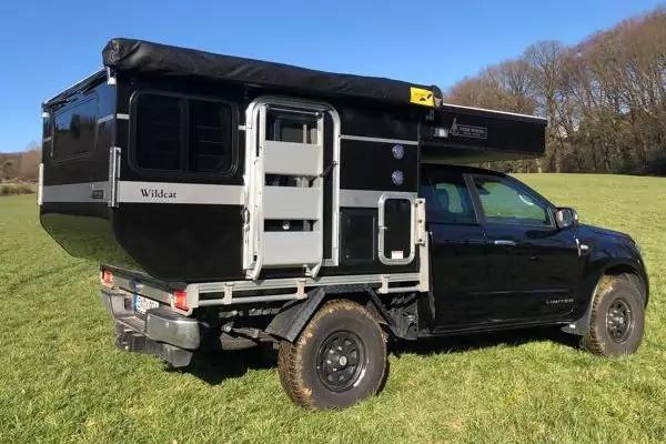 Schwarzes Pickup mit aufgesetzter Wohnkabine auf gr&uuml;nem Feld, B&auml;ume und blauer Himmel im Hintergrund