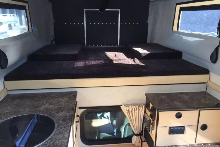 Innenraum mit erh&ouml;hter Schlafplattform und dunkler Matratze, zwei seitliche Fenster, Arbeitsplatte mit rundem Sp&uuml;lbecken, Schr&auml;nke und kleines Fenster mit Vorhang