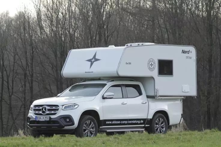 Wei&szlig;er Mercedes-Pickup mit aufgesetzter Wohnkabine auf einer Grasfl&auml;che, im Hintergrund kahle Laubb&auml;ume