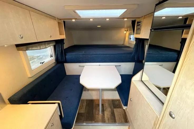 Innenraum eines Wohnmobils: Sitzb&auml;nke mit blauen Polstern, kleiner wei&szlig;er Tisch, erh&ouml;htes Bett im Heck, Dachfenster, Fenster mit dunklen Vorh&auml;ngen, hellholzfarbene Schr&auml;nke und Spiegel