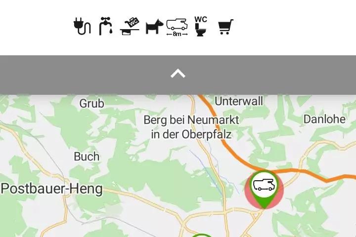 Kartenausschnitt mit Ortsnamen und einer gr&uuml;nen Markierung mit wei&szlig;em Wohnmobil‑Symbol; oben eine Reihe schwarzer Piktogramme (Stecker, Wasserhahn, Dusche, Wohnwagen, WC, Einkaufswagen)