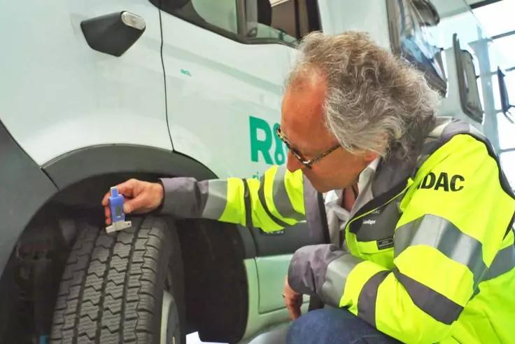Person mit grauen Haaren und gelber Warnjacke kniet neben einem Reifen eines wei&szlig;en Pannenhilfsfahrzeugs und h&auml;lt ein Messger&auml;t am Reifenprofil