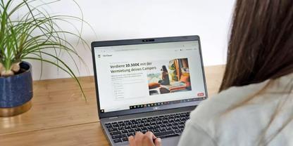 Person von hinten, H&auml;nde auf Laptop-Tastatur; Laptopbildschirm zeigt eine Webseite mit Bild eines Campers; Topfpflanze links auf holzfarbigem Schreibtisch