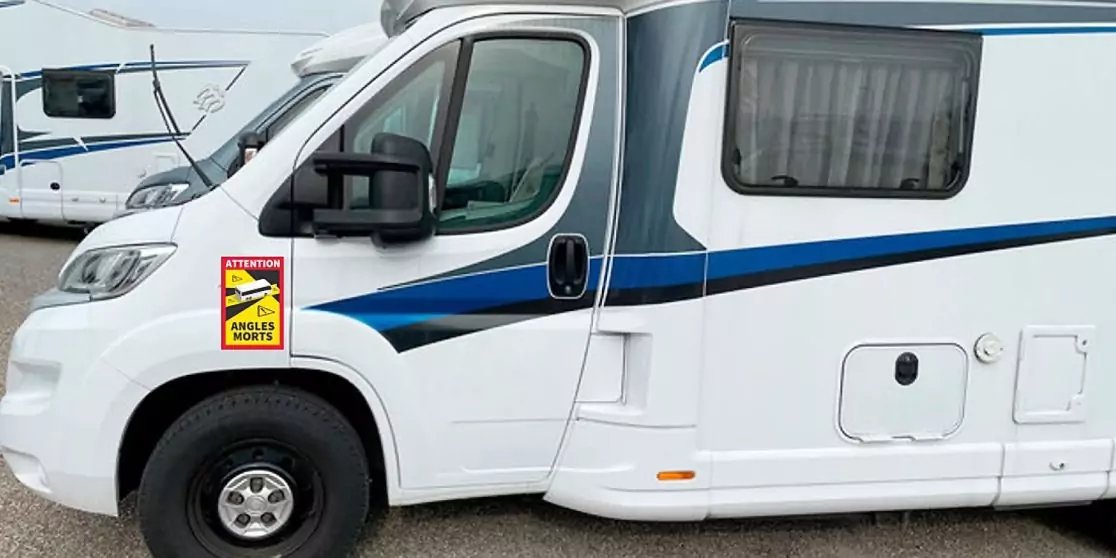 Seitenvorderansicht eines wei&szlig;en Wohnmobils mit blauem Streifen, vorderem Rad, Seitenspiegel, T&uuml;rfenster und Seitenfenster mit Vorhang, kleiner gelber Aufkleber mit schwarzem Piktogramm auf der vorderen Seitenverkleidung