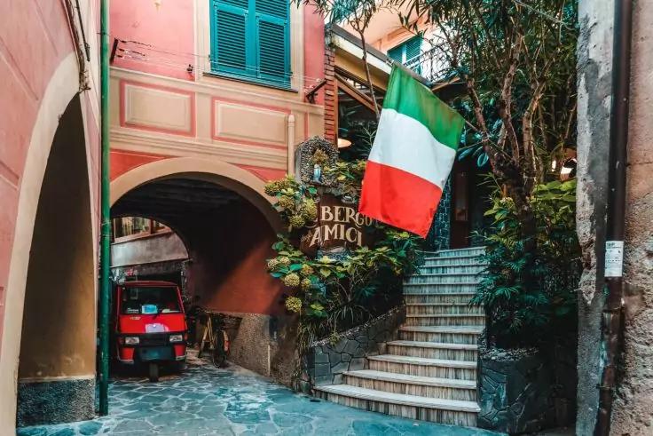 Enger gepflasterter Gassenabschnitt mit Bogen, Steintreppe, gr&uuml;nen Fensterl&auml;den, Pflanzen, h&auml;ngender gr&uuml;n-wei&szlig;-roter Flagge und kleinem roten Dreiradfahrzeug