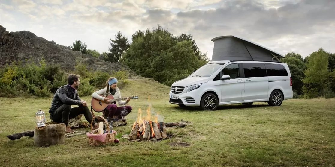 Zwei Personen sitzen am Lagerfeuer, eine spielt Gitarre; Picknickutensilien auf dem Gras; wei&szlig;er Campervan mit aufgeklapptem Aufstelldach; B&auml;ume und bew&ouml;lkter Himmel