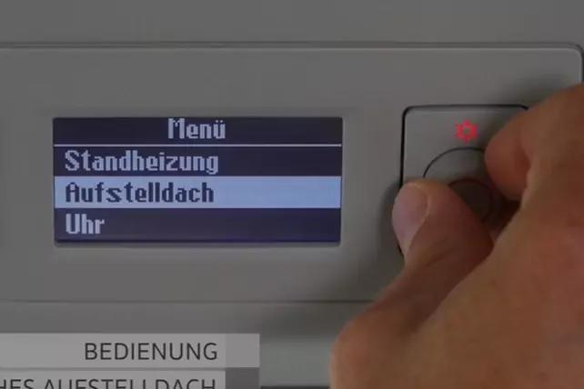 Hand dreht einen Drehknopf an einem Bedienfeld; kleines blaues Display zeigt 'Menü', 'Standheizung', 'Aufstelldach', 'Uhr'