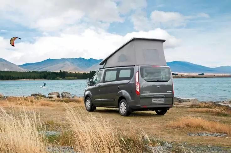 Grau lackiertes Wohnmobil mit aufgeklapptem Aufstelldach am Ufer eines Sees, im Hintergrund Berge und bewölkter Himmel, links ein bunter Kite am Himmel