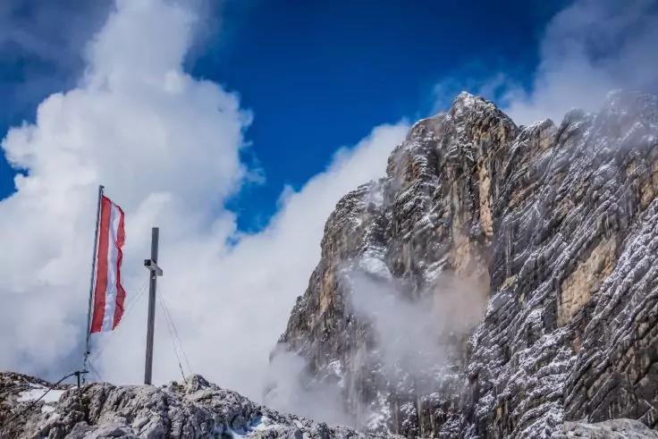 Schneebedeckter Felsgrat mit rot-wei&szlig;-roter Flagge an einem Mast mit Querbalken, steiler Fels mit Schneeflecken, Nebel- und Wolkenfetzen, blauer Himmel