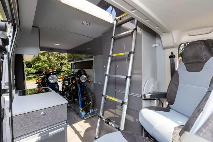 Innenraum eines Campers mit zwei Motorrädern im Heck, einer Leiter zum Hochbett, grauen Schränken, einem drehbaren Sitz und einem Seitenfenster mit Bäumen