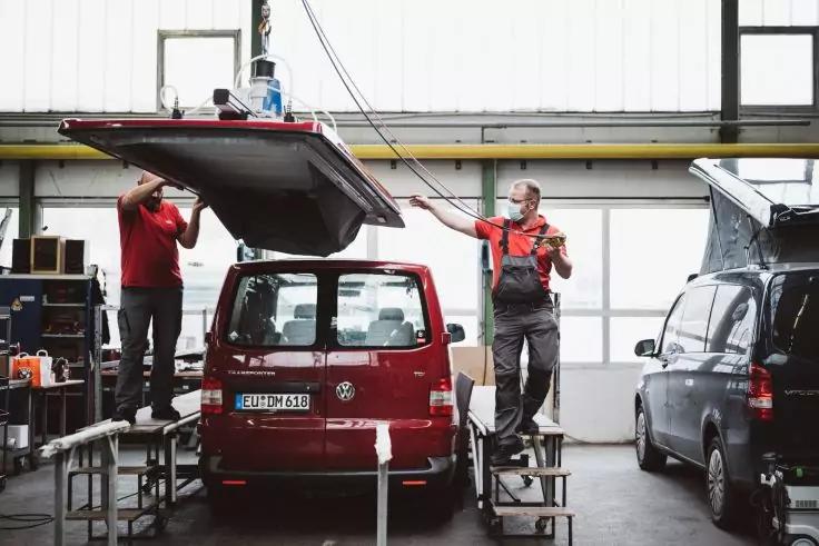 Zwei Personen heben eine Dachbox auf das Dach eines roten Kleinbusses in einer Werkstatt