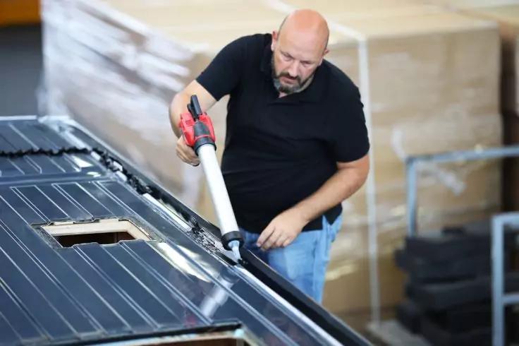 Glatzköpfiger Mann in schwarzem T‑Shirt hält eine Kartuschenpistole und trägt Dichtstoff auf ein dunkles Metall-Dachpaneel auf; Kartonpaletten im Hintergrund
