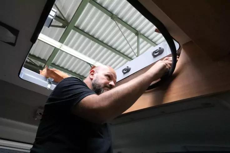 Ein Mann mit Bart greift durch eine offene Dachluke im Innenraum eines Fahrzeugs; außen sind Metallträger sichtbar