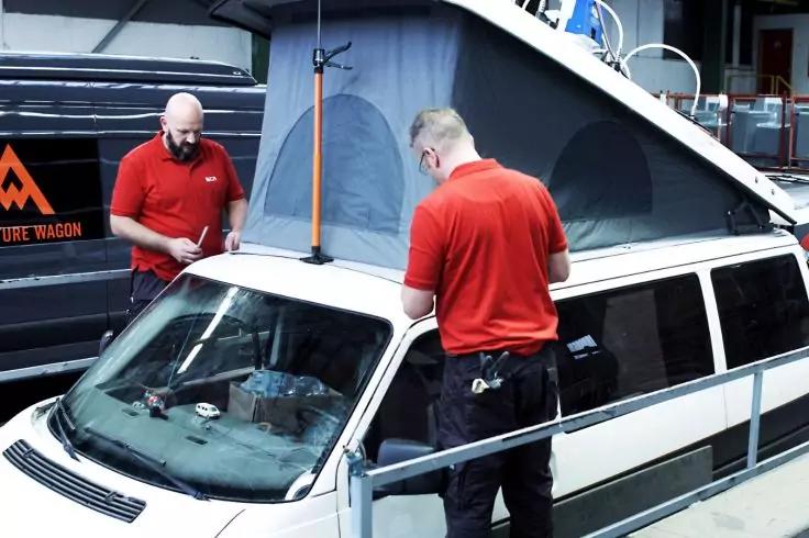 Zwei Personen in roten Poloshirts stehen an einem weißen Fahrzeug mit aufgeklapptem Aufstelldach; im Hintergrund ein weiteres Fahrzeug, im Vordergrund ein Metallgeländer