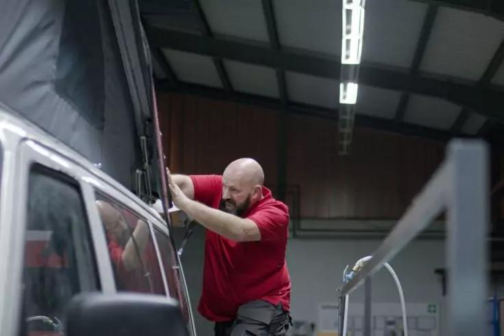 Person mit Glatze und Bart in rotem T‑Shirt hält eine Plane an der Seite eines Fahrzeugs; Deckenbeleuchtung und ein Geländer im Innenraum sichtbar