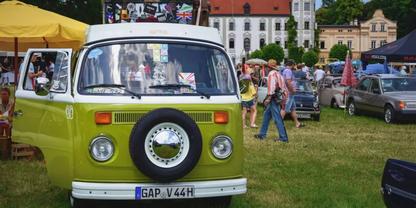 Vorderansicht eines gr&uuml;nen VW-Campers mit aufgesetztem Ersatzrad, im Hintergrund Menschen, Zelte und ein Geb&auml;ude