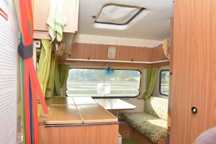 Holzvertäfelte Schränke, zusammengeklappter Tisch, grün gemustertes Polster, grüne Vorhänge, Fenster mit Blick auf Straße und Bäume, Deckenlüfter, grünes Handtuch, kleiner blauer Clipventilator, rot‑orange gestreifter Gurt