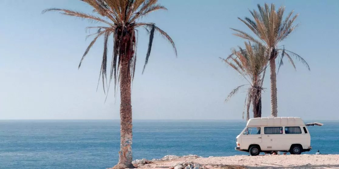 Wei&szlig;er Van auf felsigem Ufer zwischen zwei Palmen, im Hintergrund blaues Meer und klarer Himmel