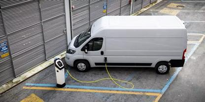 Wei&szlig;er Lieferwagen auf einem Parkplatz neben Metall-Rolltoren, &uuml;ber ein Kabel mit einer Ladestation verbunden