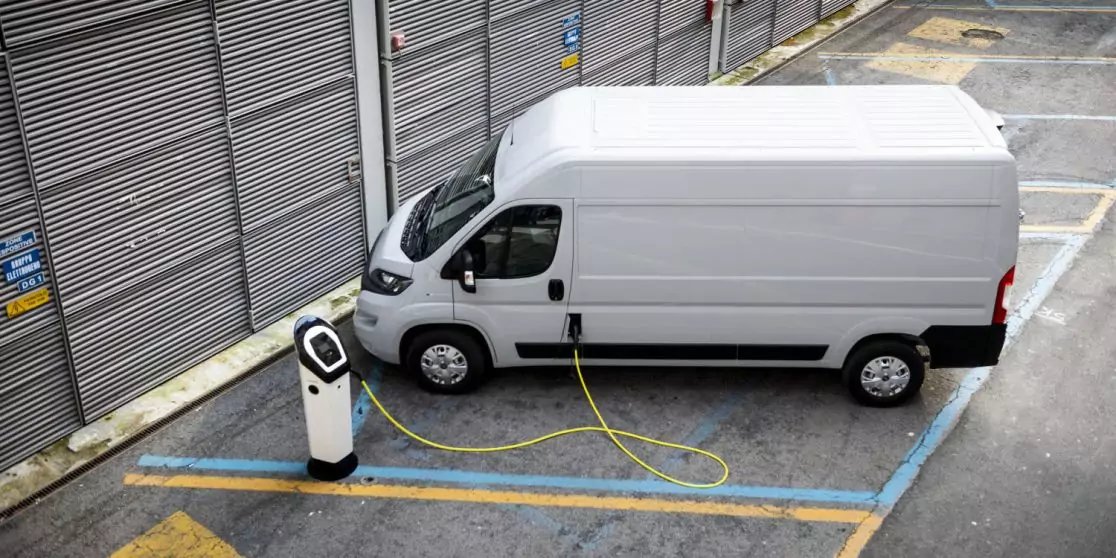 Wei&szlig;er Lieferwagen auf einem Parkplatz neben Metall-Rolltoren, &uuml;ber ein Kabel mit einer Ladestation verbunden