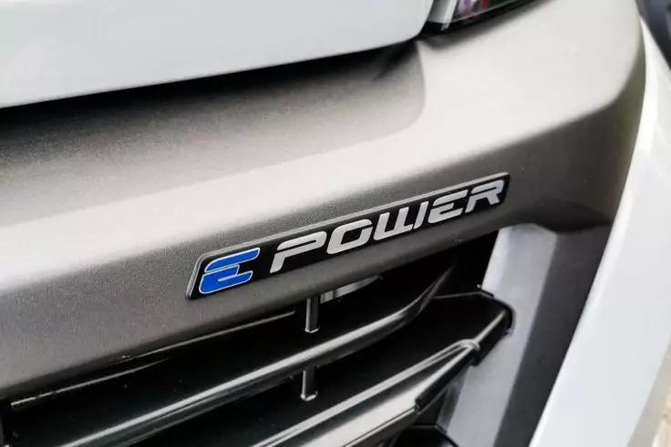 Nahaufnahme der Fahrzeugfront: silberner Sto&szlig;f&auml;nger, schwarzer K&uuml;hlergrill mit horizontalen Lamellen, Emblem "E-POWER" mit blauem E, Teil eines Scheinwerfers