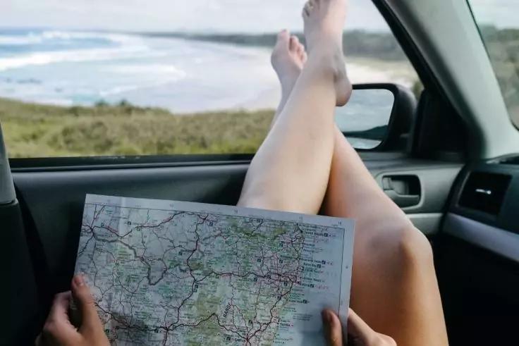 Beine einer Person auf dem Armaturenbrett, H&auml;nde halten eine aufgeklappte Stra&szlig;enkarte; durch das Seitenfenster Strand, D&uuml;nen und Meer