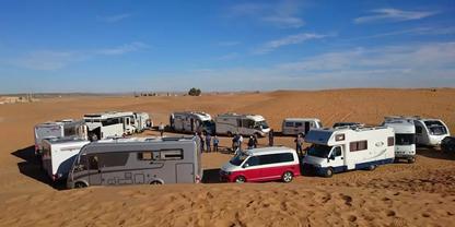 Mehrere Wohnmobile, ein rot-wei&szlig;es Auto und Anh&auml;nger auf Sandd&uuml;nen; Personen zwischen den Fahrzeugen, blauer Himmel mit vereinzelten Wolken