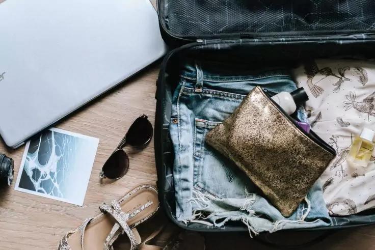 Offener Koffer mit zerrissener Jeans, goldfarbener Kosmetiktasche, Sonnenbrille, Sandalen, teilweise sichtbarem Laptop, Fotoabzug und kleinen Flaschen auf Holzboden