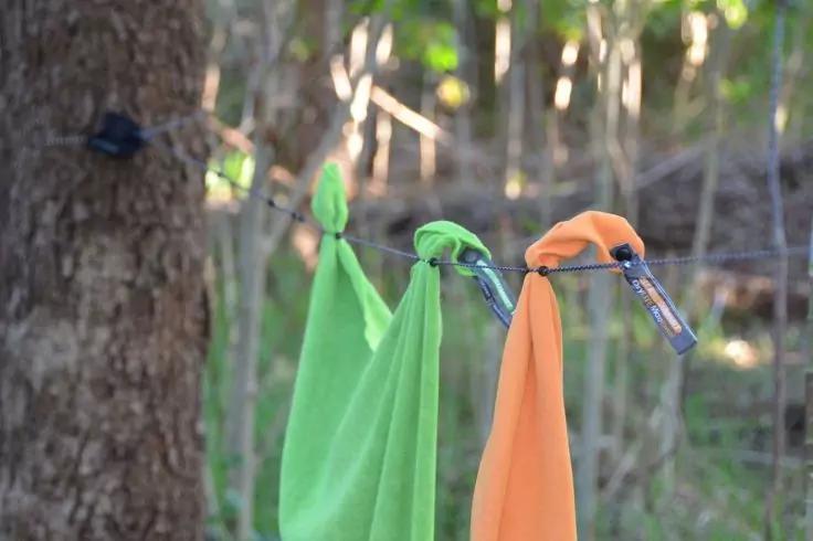 Gr&uuml;nes und oranges Tuch h&auml;ngen an einem d&uuml;nnen Seil, jeweils mit Metallkarabinern befestigt, unscharfer Waldhintergrund mit B&auml;umen und Zweigen