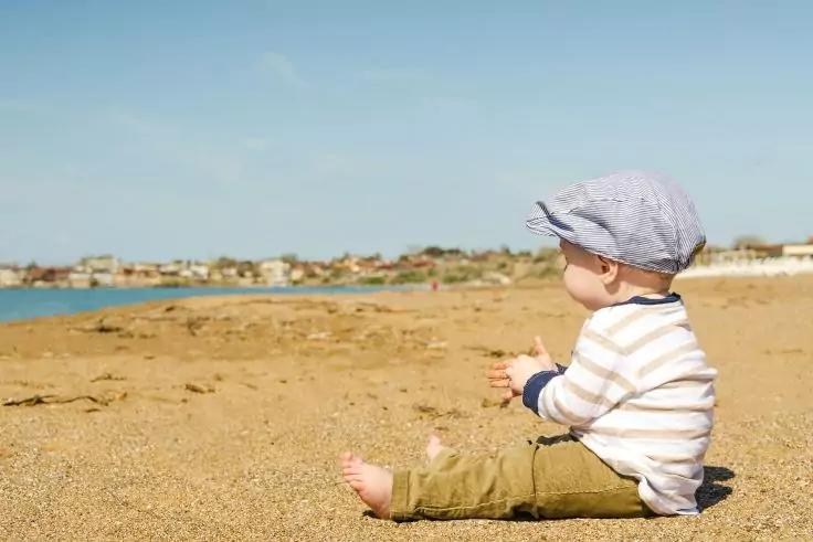 Kleinkind sitzt seitlich auf einem Sandstrand, tr&auml;gt gestreiftes Langarmshirt, olivgr&uuml;ne Hose und helle M&uuml;tze, blickt zum Wasser