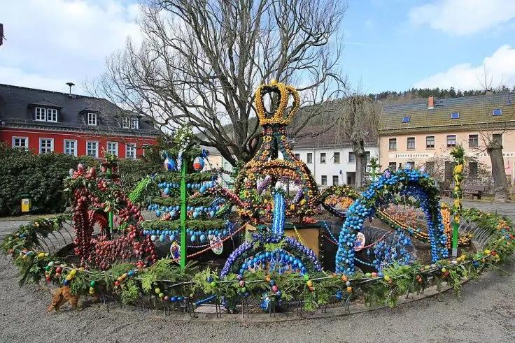 Runde Installation mit mehreren bogenf&ouml;rmigen Gestellen und einer kr&ouml;nchenartigen Spitze, dicht mit bunten Eiern und gr&uuml;nen Zweigen geschm&uuml;ckt; kahler Baum und mehrst&ouml;ckige Geb&auml;ude im Hintergrund, blauer Himmel mit Wolken