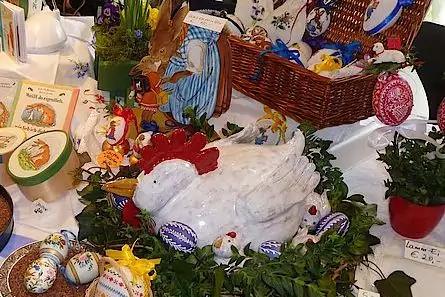 Wei&szlig;e Keramikhenne auf einem Kranz aus gr&uuml;nen Bl&auml;ttern, umgeben von bemalten Eiern, kleinen K&uuml;ken- und Hasenfiguren sowie einem geflochtenen Korb mit weiteren Eiern