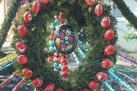 Gr&uuml;ner Kranz aus Tannenzweigen mit roten, bemalten Eiern; mittig ein schwarzes Ei mit Blumenmuster; im Hintergrund mehrfarbige Perlen- und Eiergirlanden